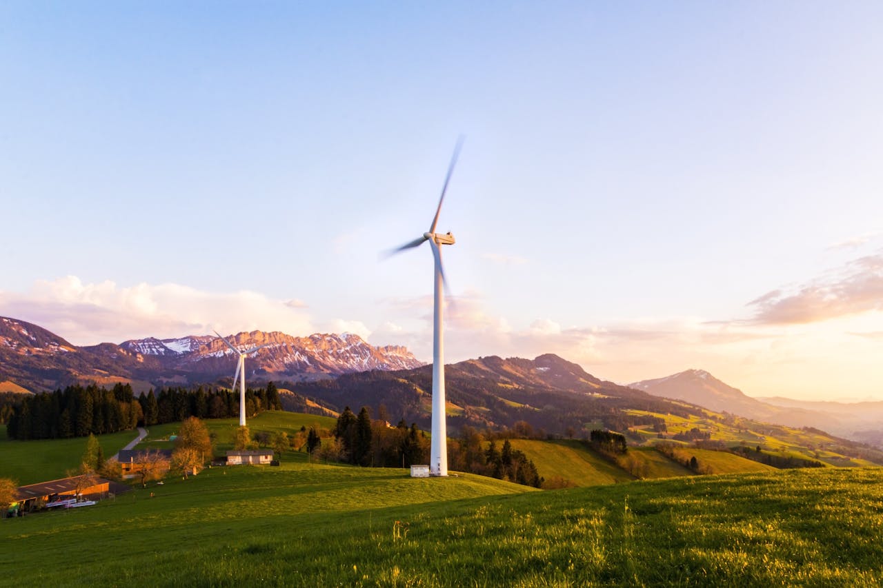 about-01 A wind turbine stands tall against a mountainous backdrop under a vibrant sky at sunset.